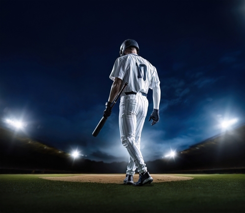 shutterstock_2431528815.jpg A baseball player standing on the field at night, holding a bat