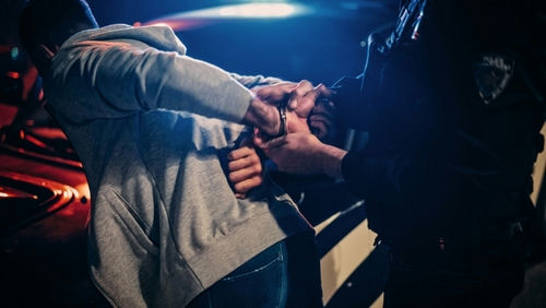shutterstock2232056691-1jpg Person being handcuffed by an officer beside a police car at night