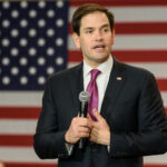 Rubio vs. Vance: The Evangelical Mystery… A politician speaking into a microphone in front of an American flag