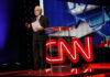 CNN Brawl Explodes Over Musk Label news anchor stands on stage beside a clear podium