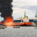 Drone Barrage Upends Crimea’s Backbone A large ship on fire with black smoke rising, surrounded by smaller fishing boats in a harbor
