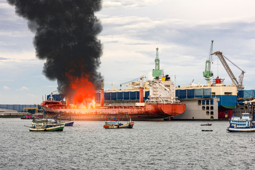Large,General,Cargo,Ship,For,Logistic,Import,Export,Goods,And A large ship on fire with black smoke rising, surrounded by smaller fishing boats in a harbor