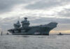 Aircraft carrier with tugboats on a cloudy sea.