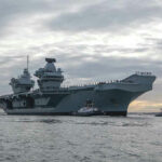 Aircraft carrier with tugboats on a cloudy sea.