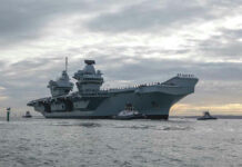 Legal Loophole Keeps ‘Dead’ Carrier Alive Aircraft carrier with tugboats on a cloudy sea.