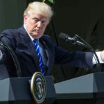 Man speaking at a podium, arms extended outward.