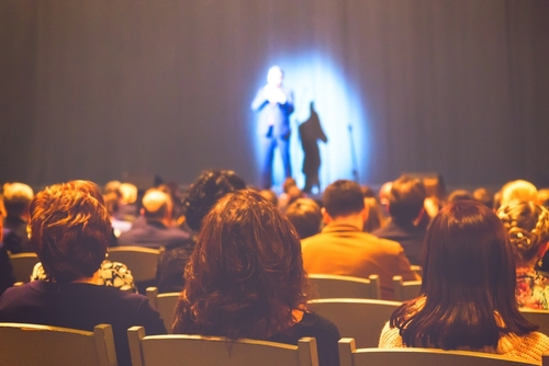 Man,Appears,On,Stage,In,Theater,With,A,Lot,Of Audience watching a performance on stage with dramatic lighting