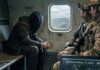 A hooded figure sits quietly in a helicopter with soldiers, overlooking a city skyline.