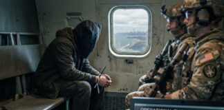 A hooded figure sits quietly in a helicopter with soldiers, overlooking a city skyline.
