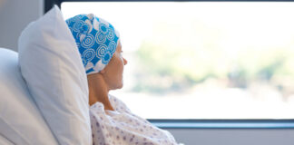A female patient in a hospital bed wearing a blue patterned headscarf, looking out a window