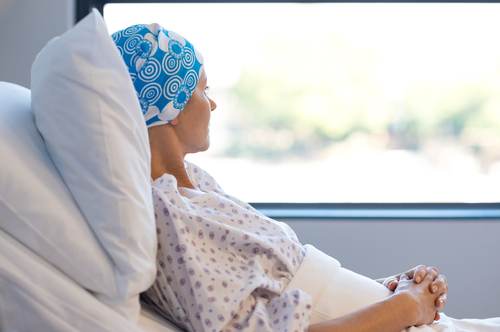 A female patient in a hospital bed wearing a blue patterned headscarf, looking out a window