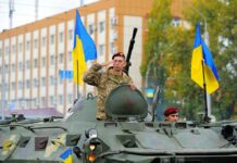 Soldier saluting from armored vehicle with Ukrainian flags.